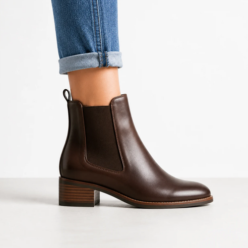 Une femme porte des bottines Chelsea marron en cuir avec des inserts élastiques et un talon bloc, associées à un jean retroussé dans un studio.