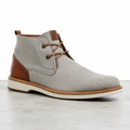 Une bottine Chukka grise en tissu avec des détails en cuir marron et des lacets, posée sur un sol clair dans un studio.   