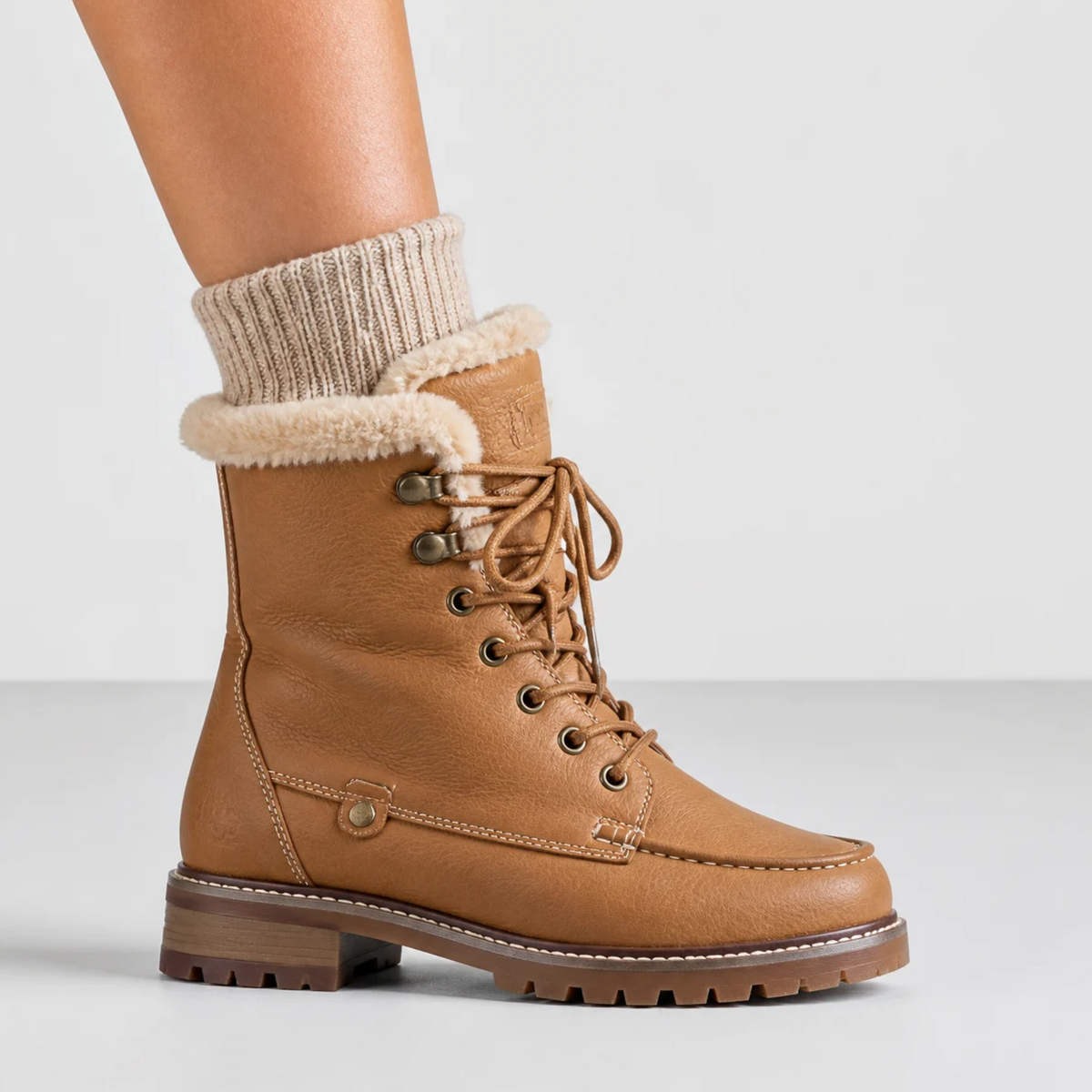 Une femme porte des bottines d'hiver marron en cuir fourrées avec une doublure en fausse fourrure, des lacets et une semelle crantée dans un studio.