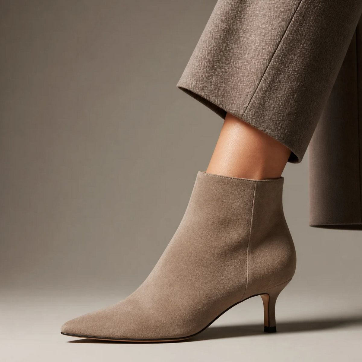 Une femme porte des bottines beiges en suédine avec un bout pointu et un talon fin, associées à un pantalon habillé dans un studio.