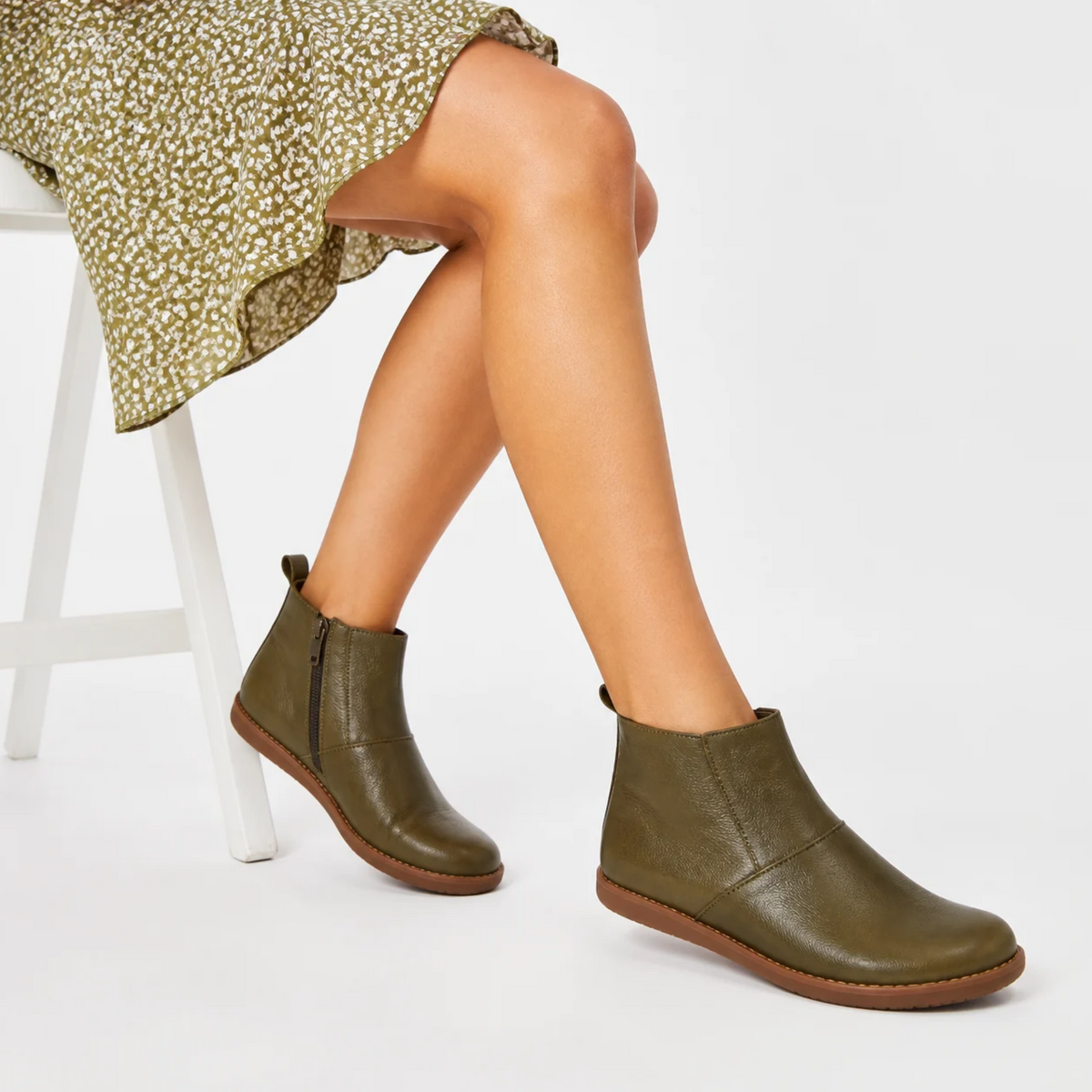 Une femme assise porte des bottines vert olive en cuir avec une fermeture éclair latérale, associées à une robe fleurie.