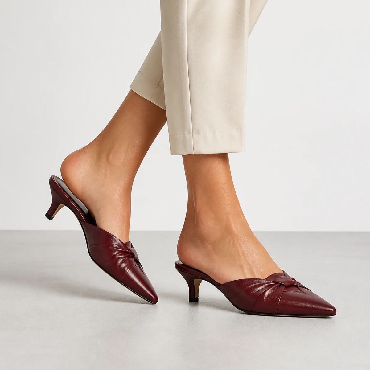 Une femme porte des mules bordeaux en cuir avec un détail noué, un bout pointu et un petit talon, associées à un pantalon clair dans un studio.