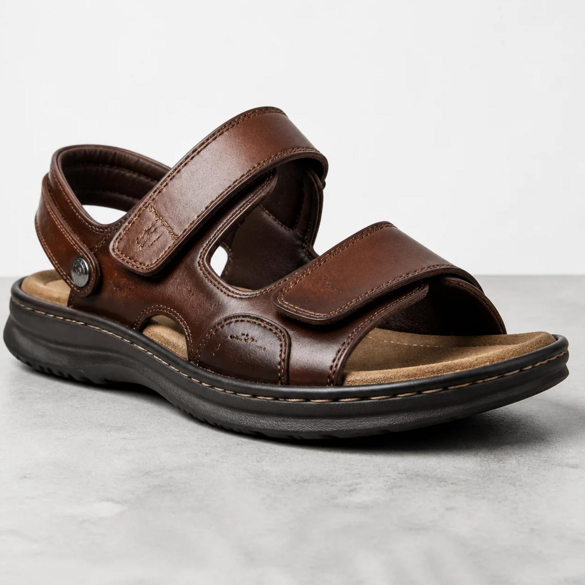 Une sandale marron en cuir pour homme avec des brides ajustables, posée sur un sol clair dans un studio.   