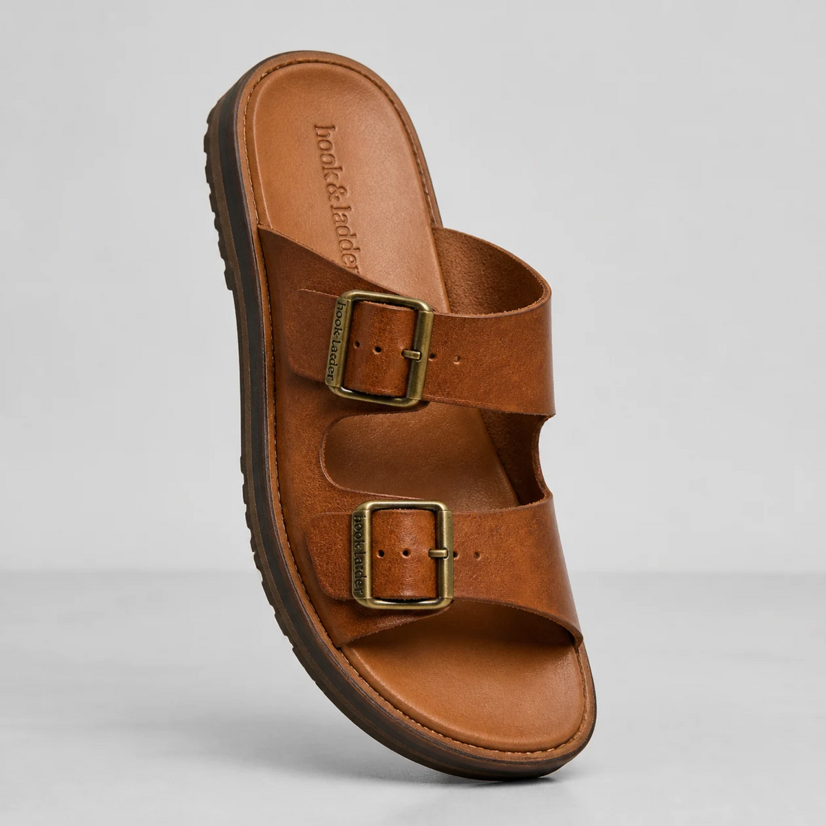 Une sandale plate marron en cuir pour homme avec deux larges brides à boucles métalliques, présentée en studio sur un fond neutre.   